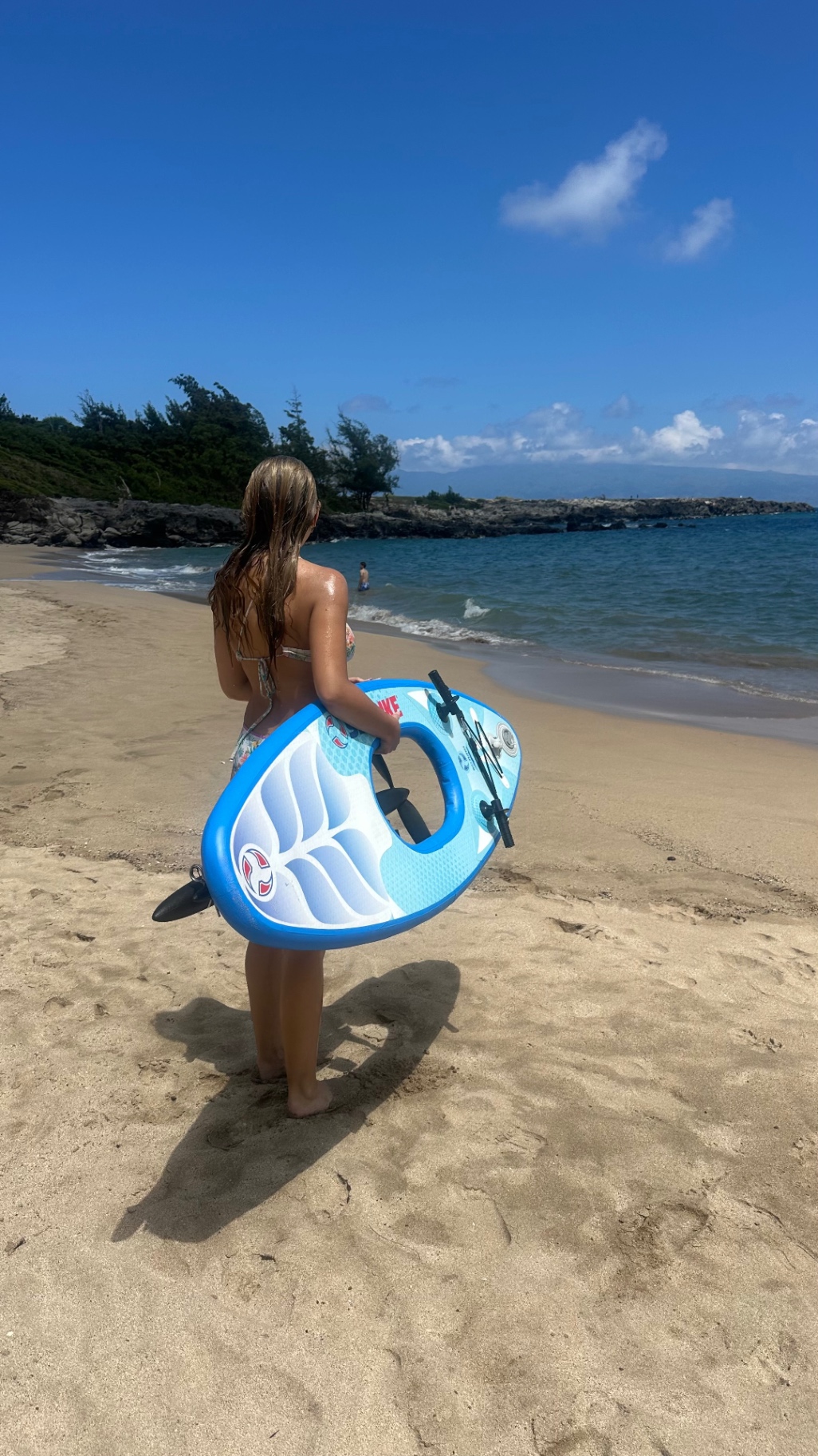 Guest carrying a lightweight Maui Sea Board snorkeling board along the wet sand at the shoreline, ocean and blue sky in the background